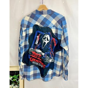 Może przedstawiać: Niebiesko-biała koszula w kratę z flaneli z grafiką Ghostface Movie Club na plecach. Grafika przedstawia postać Ghostface trzymającą nóż, ze słowami "Ghostface Movie Club" w kolorze czerwonym i białym.