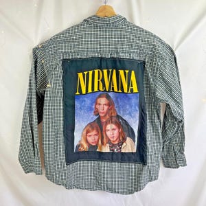 May include: Green and white plaid button-down shirt with a Nirvana band graphic on the back. The graphic features the band's name in yellow letters above a photo of the band members. The shirt has long sleeves and a classic collar.