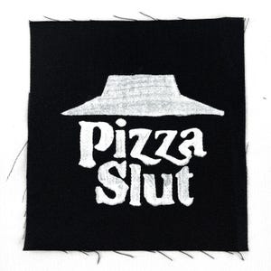 May include: Black square patch with white embroidered text and a hat graphic. The text reads "Pizza Slut" in a bold, stylized font. The hat graphic is above the text, and the patch has raw edges.