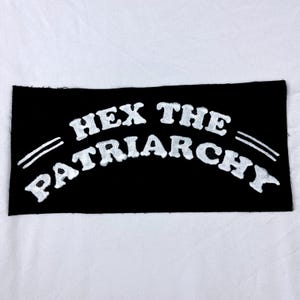 May include: Black patch with the words "HEX THE PATRIARCHY" in white, arched lettering. Two white parallel lines are on the left side. The patch is rectangular and has a slightly frayed edge.