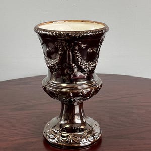 May include: A brown ceramic urn with a pedestal base. The urn features ornate relief detailing, including floral and garland motifs. The rim is a lighter cream color. The urn is displayed on a dark wood surface.