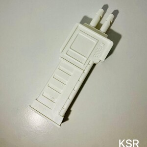 May include: A white plastic toy gun accessory with a rectangular body and two protruding cylinders. The accessory has a detailed design with rectangular and textured elements. The letters "KSR" are visible in the bottom right corner.