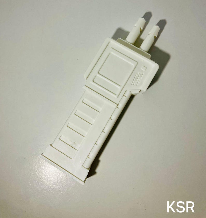 May include: A white plastic toy gun accessory with a rectangular body and two protruding cylinders. The accessory has a detailed design with rectangular and textured elements. The letters "KSR" are visible in the bottom right corner.