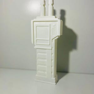 May include: A white, futuristic-looking sculpture with a rectangular base and a series of horizontal lines. The top section features a square screen and two vertical cylinders. The letters "KSR" are visible at the bottom.