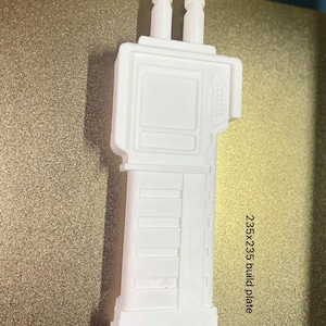 May include: A white 3D-printed object with a rectangular body and two vertical protrusions. The object has a detailed design with rectangular and square features. The text "235x235 build plate" is visible. The letters "KSR" are also visible.