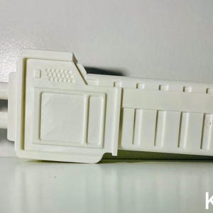 May include: A white, plastic, futuristic-looking object with a rectangular body and two cylindrical protrusions. The object has a series of vertical lines on one side and a textured surface on the other. The letters "KSR" are visible.