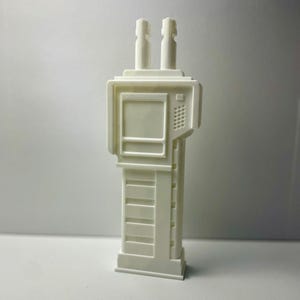 May include: A white, 3D-printed model of a futuristic handheld device. The device has two cylindrical antennas, a rectangular screen, and a keypad. The base is rectangular with horizontal lines. The letters "KSR" are visible at the bottom.