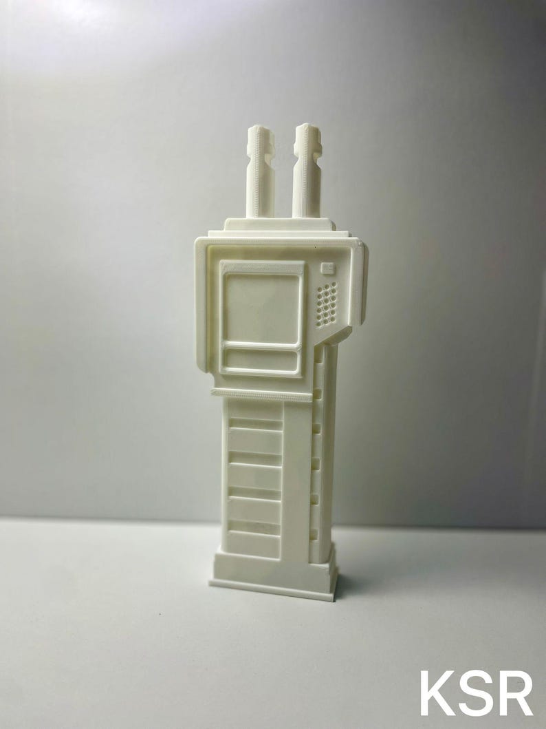 May include: A white, 3D-printed model of a futuristic handheld device. The device has two cylindrical antennas, a rectangular screen, and a keypad. The base is rectangular with horizontal lines. The letters "KSR" are visible at the bottom.