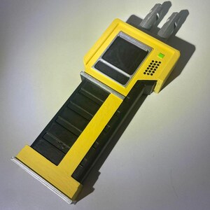 May include: A yellow and black device with a rectangular screen and two gray prongs. The device has a long, black handle with yellow accents and a small green light.