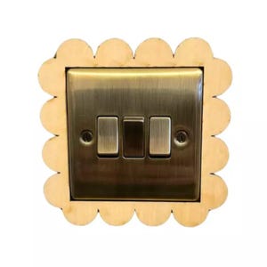 May include: A decorative light switch plate with a scalloped wooden frame. The plate is a dark brass colour and features three rectangular switches. The wooden frame is a light tan colour.