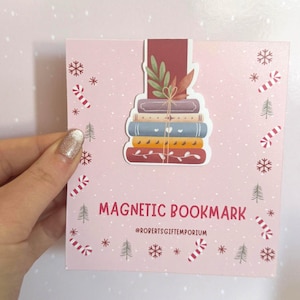 May include: A pink magnetic bookmark with a stack of colourful books and a leaf design. The text "MAGNETIC BOOKMARK" is displayed on the card, along with the shop's name. The card is decorated with snowflakes, candy canes, and Christmas trees, evoking a festive theme.