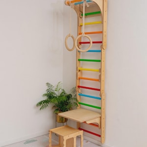 Pode incluir: Uma parede de escalada de madeira com degraus coloridos do arco-íris e uma mesa dobrável. A parede é montada em uma parede branca e inclui dois anéis de madeira pendurados no topo.