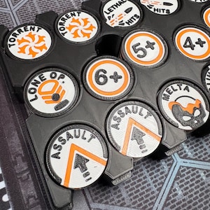 Puede incluir: Una bandeja negra contiene varias fichas de juego blancas y negras con detalles en naranja. Las fichas presentan varios símbolos y texto, incluyendo "Torrent", "Lethal Hits" y números como "6+" y "5+." La bandeja está diseñada para organizar las fichas.
