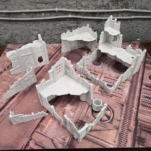 Könnte beinhalten: Eine Sammlung grauer, miniaturisierter, zerstörter Gebäudemodelle. Die Modelle weisen detaillierte architektonische Elemente auf, darunter Wände, Böden und zerbrochene Abschnitte. Die Teile sind auf einer braunen und grauen Oberfläche angeordnet, was auf ein Tabletop-Spiel hindeutet.