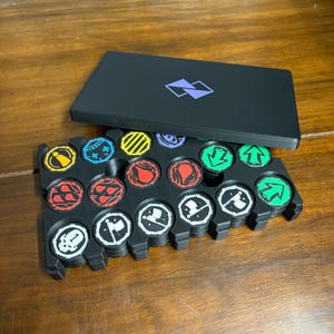 Könnte beinhalten: Ein schwarzes Spielbrett mit einem Tablett voller bunter Spielsteine. Die Spielsteine haben verschiedene Symbole und Designs in Rot, Gelb, Blau und Grün. Eine schwarze rechteckige Box mit einem lila Logo liegt obenauf.