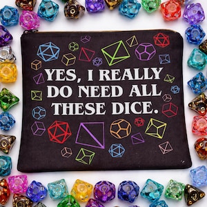 May include: Black zippered pouch with the text "YES, I REALLY DO NEED ALL THESE DICE." surrounded by colorful geometric shapes. The pouch is framed by a collection of various colorful dice.