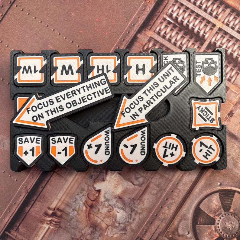 May include: Black and orange game tokens and markers. Includes tokens with text such as "Wound", "Save", and "Hit". Also includes arrows with the text "Focus Everything on this Objective" and "Focus this Unit in Particular".