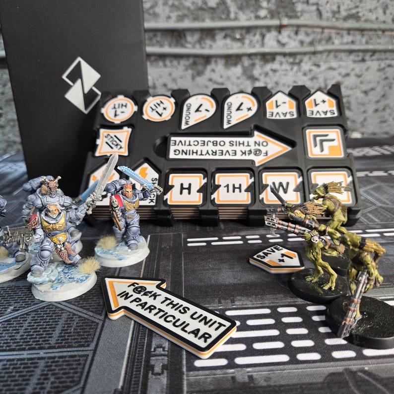 May include: A tabletop gaming scene featuring miniature figures and game tokens. The figures are painted in various colors and posed in action. Tokens include phrases like "F@ck this unit in particular" and "F@ck everything on this objective".