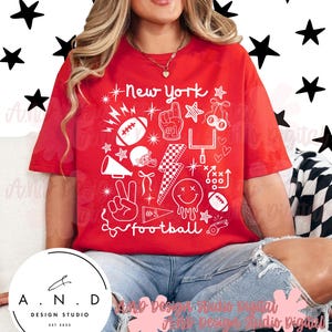 Puede incluir: Una camiseta roja con un diseño gráfico blanco que presenta el texto "New York" e iconos relacionados con el fútbol. El diseño incluye un balón de fútbol, un casco y una cara sonriente. La camiseta la lleva una persona.