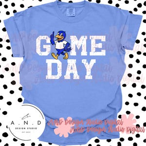 May include: A light blue t-shirt with the words "GAME DAY" in distressed white lettering. A cartoon bird in blue, white, and yellow is pointing upwards. The shirt has a crew neck and short sleeves. The design studio logo is at the bottom.