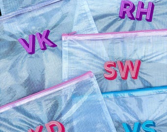 May include: A collection of clear mesh zipper pouches with various embroidered initials in different colors. The pouches have colored zippers and initials in shades of blue, pink, and purple. The initials include MB, VK, RH, SW, KD, VS, and BK.