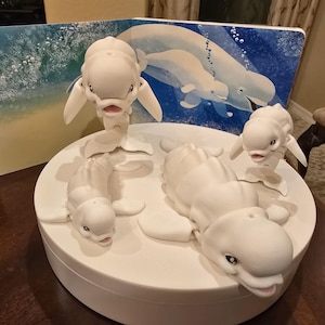 May include: Four white plastic figurines of beluga whales on a white circular base. The whales are in various poses, with one standing upright and the others lying down.