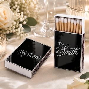 May include: Two rectangular matchboxes with a glossy black finish and white text. One box is upright, filled with wooden matches with white tips, displaying the text The Smith. The second box lies flat, featuring the text July 27, 2026.