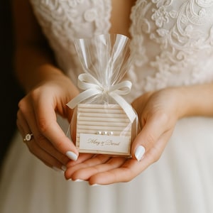 May include: A small, rectangular wedding favour wrapped in clear cellophane and tied with a white ribbon. The favour is a light beige colour with a small label that reads "Mary & Derek" and includes three small pearls.