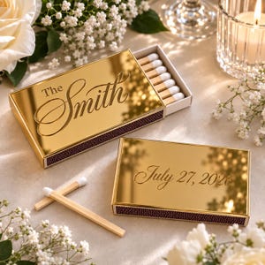 May include: Two rectangular matchboxes with a reflective gold finish. One box is open, revealing wooden matches with white tips, and features the text The Smith in script. The second closed box displays the text July 27, 2026 in script.