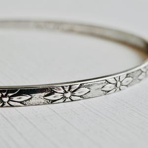 Op de afbeelding: Een zilverkleurige armband met een bloemmotief. De armband heeft een reeks in reliëf aangebrachte bloemen rond de band. De bloemen hebben gedetailleerde bloemblaadjes en een centraal ontwerp. De armband wordt getoond op een wit oppervlak.