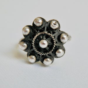 Könnte beinhalten: Ein silberner Ring mit floralem Design. Der Ring hat eine zentrale Kugel, die von einem gedrehten Seilmuster umgeben ist, das von einem Ring aus silbernen Kugeln umschlossen wird. Der Ring ist sichtbar, und das Design ist vor einem weißen Hintergrund platziert.