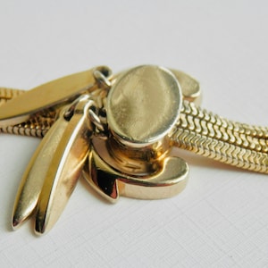 May include: A gold-toned vintage necklace clasp with a unique design. The clasp features an oval center piece, decorative curved elements, and two elongated, leaf-shaped pendants. The chain is a flat, woven style.