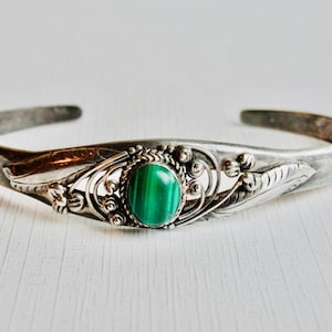 May include: Silver cuff bracelet with a central oval malachite stone in a vivid green hue. The bracelet is adorned with detailed floral and leaf patterns, enhancing its decorative appeal.