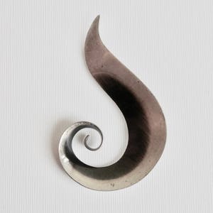 Op de afbeelding: Een metalen broche in de vorm van een gestileerde vlam of een golvende curve die bovenaan uitloopt in een punt en onderaan krult in een strakke spiraal. Het oppervlak heeft een geborstelde, donker zilverkleurige afwerking met zichtbare textuur.