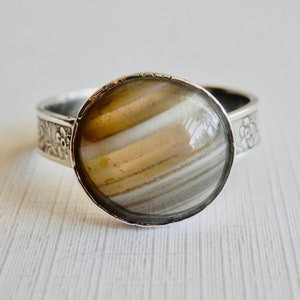 May include: A silver ring featuring a round, cabochon-style stone with horizontal bands of tan, white, and gray. The metal band displays a detailed, embossed floral pattern along its sides, framing the smooth, polished stone centerpiece.