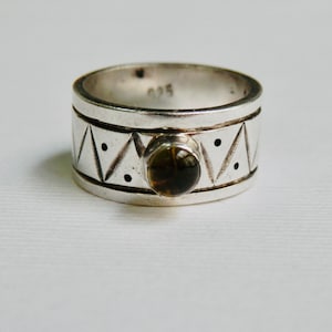 May include: A silver ring with a geometric design and a dark brown gemstone. The ring has a wide band with etched triangles and dots. The gemstone is round and set in a bezel. The ring is a size 9.25.