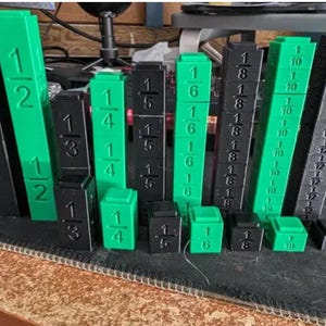 May include: A set of black and green fraction blocks. The blocks are made of plastic and have different sizes and shapes. The blocks are labeled with fractions, such as 1/2, 1/3, 1/4, 1/5, 1/6, 1/8, 1/10, and 1/12.