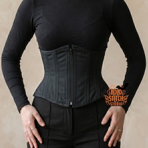 May include: A black corset with a front zipper is worn over a black long-sleeve shirt. The corset has vertical seams and a structured design. The model is wearing black pants. The Bold Stitch Studio logo is visible on the right side.