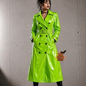 Könnte beinhalten: Ein leuchtend lindgrüner Trenchcoat aus glänzendem, lackartigem Material. Der Mantel hat eine zweireihige Vorderseite mit schwarzen Knöpfen, einen passenden Gürtel mit Schnalle, breite Revers und lange Ärmel. Ein Logo zeigt BOLD STITCH STUDIO.