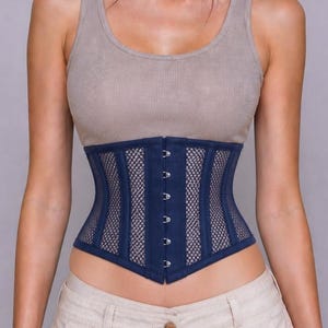 May include: A navy blue corset with a mesh design is worn over a beige tank top. The corset has a structured shape with vertical lines and a row of hooks down the front. The model is also wearing beige pants.