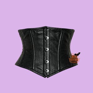 May include: A black corset featuring a smooth, matte finish with vertical paneling and a front closure consisting of four silver-toned metal clasps. The garment displays a logo on the lower right side with the text Bold Stitch Studio.