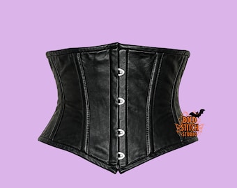 May include: A black corset featuring a smooth, matte finish with vertical paneling and a front closure consisting of four silver-toned metal clasps. The garment displays a logo on the lower right side with the text Bold Stitch Studio.