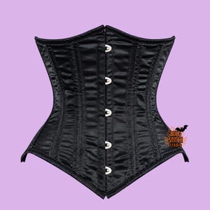 May include: A black satin corset featuring a front busk closure with five silver-toned metal clasps. The garment has vertical boning channels throughout the bodice and a pointed hemline. A small orange logo reads Bold Stitch Studio.