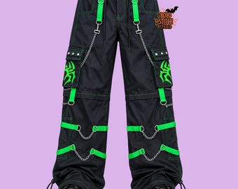 May include: Black cargo pants featuring bright green contrast stitching, neon green spider graphics on the side pockets, and silver metal chains attached to green fabric straps. The pants include multiple pockets and a Bold Stitch Studio logo.