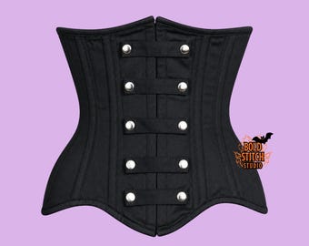 May include: A black corset featuring vertical paneling and five horizontal fabric straps across the center, each secured with a silver-toned metal snap button. The text BOLD STITCH STUDIO with a small bat graphic is visible on the lower right side.