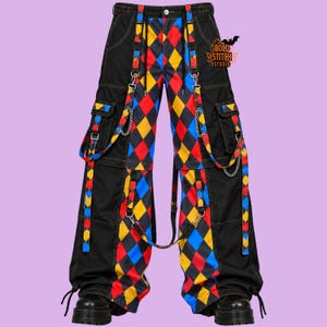 May include: Black cargo trousers featuring a vibrant red, yellow, and blue diamond harlequin pattern on the front panels. The trousers include attached straps, silver-tone metal chains, side cargo pockets, and a logo reading Bold Stitch Studio.