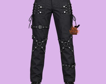 May include: Black trousers featuring side lacing, multiple adjustable straps with silver-tone metal buckles, and decorative cross-straps with metal rings on the thighs. The Bold Stitch Studio logo is visible on the right side of the trousers.