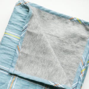 May include: A folded, quilted blanket with a light blue plaid pattern on the exterior and a gray interior. The plaid design features white, yellow, and orange stripes. The blanket is made of soft fabric.