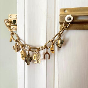 May include: Gold-coloured door chain with decorative charms. Charms include cacti, a cowboy boot, a horseshoe, and a desert landscape. The chain is attached to a gold-coloured door latch on a white door.