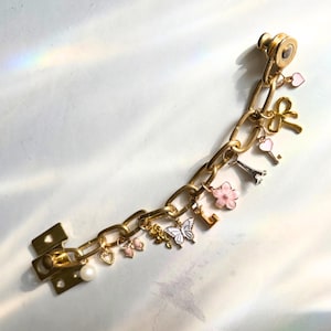 May include: Gold-coloured chain bracelet with assorted charms, including a butterfly, Eiffel Tower, hearts, and a flower. The bracelet has a clasp closure and a pearl accent. The charms are in shades of pink, white, and gold.